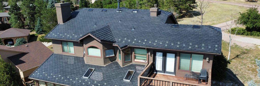 New Roof Colorado Springs – wide Aerial view of a large two-story house with a dark shingle roof, multiple chimneys, skylights, and a wooden deck surrounded by trees.