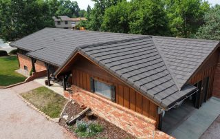 Aerial view of a single-story brick and wood house with a dark gray Campbell Roofing stone coated steel tile roof, surrounded by trees and a gravel driveway.